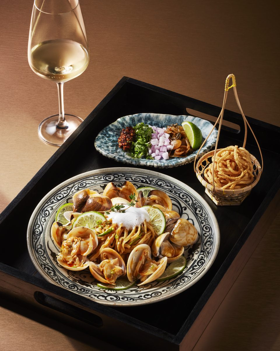 A dark tray set with noodles and shellfish, small dishes and a glass of white wine